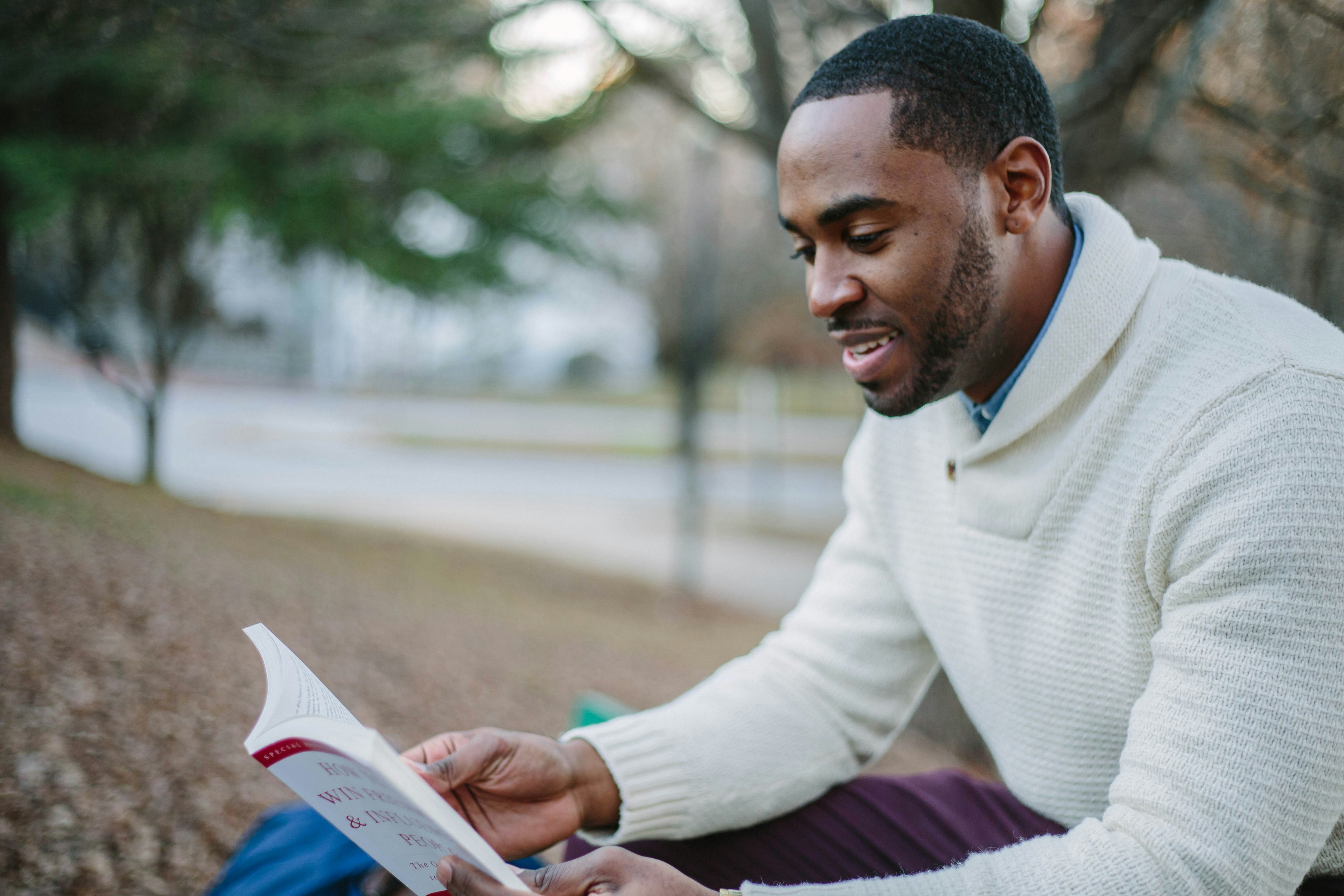 A man in his 20s sits reading a book outdoors. He wears a white turtleneck. In the background a river and trees are blurred.