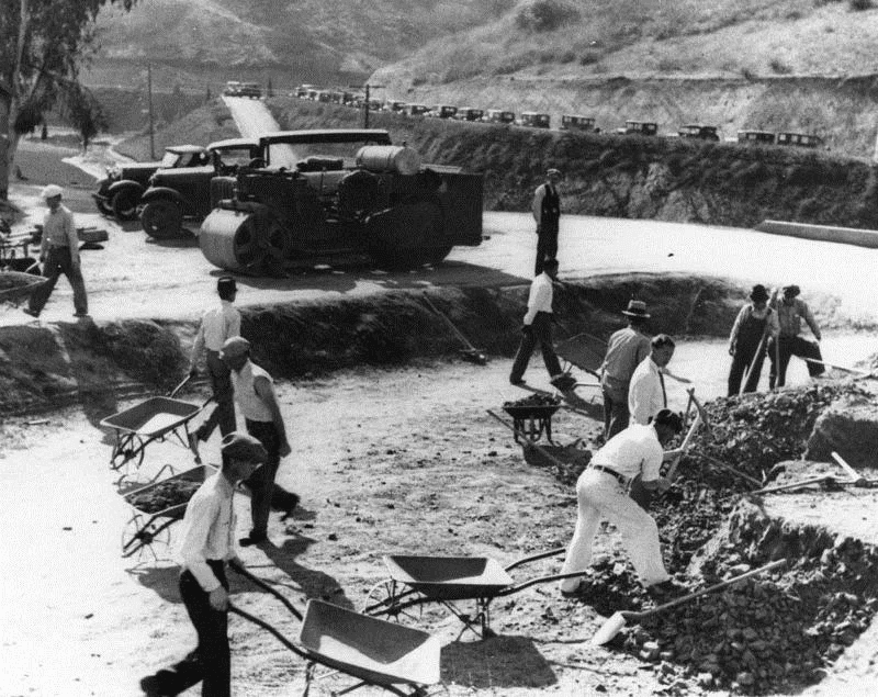 black and white photo of a contructions site with a team of worker digging a foundation with hand tools and wheel barrows with a Ford model T and an old steam roller parked in the background
