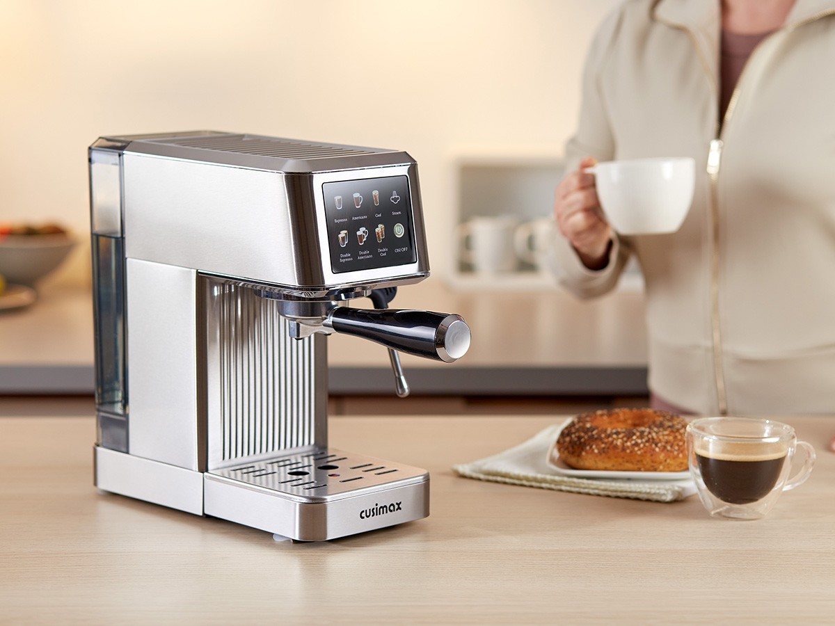 A modern stainless steel Cusimax espresso machine sits on a wooden kitchen counter, with a person holding a white coffee cup in the background, next to a plate with a pastry and a glass of espresso.