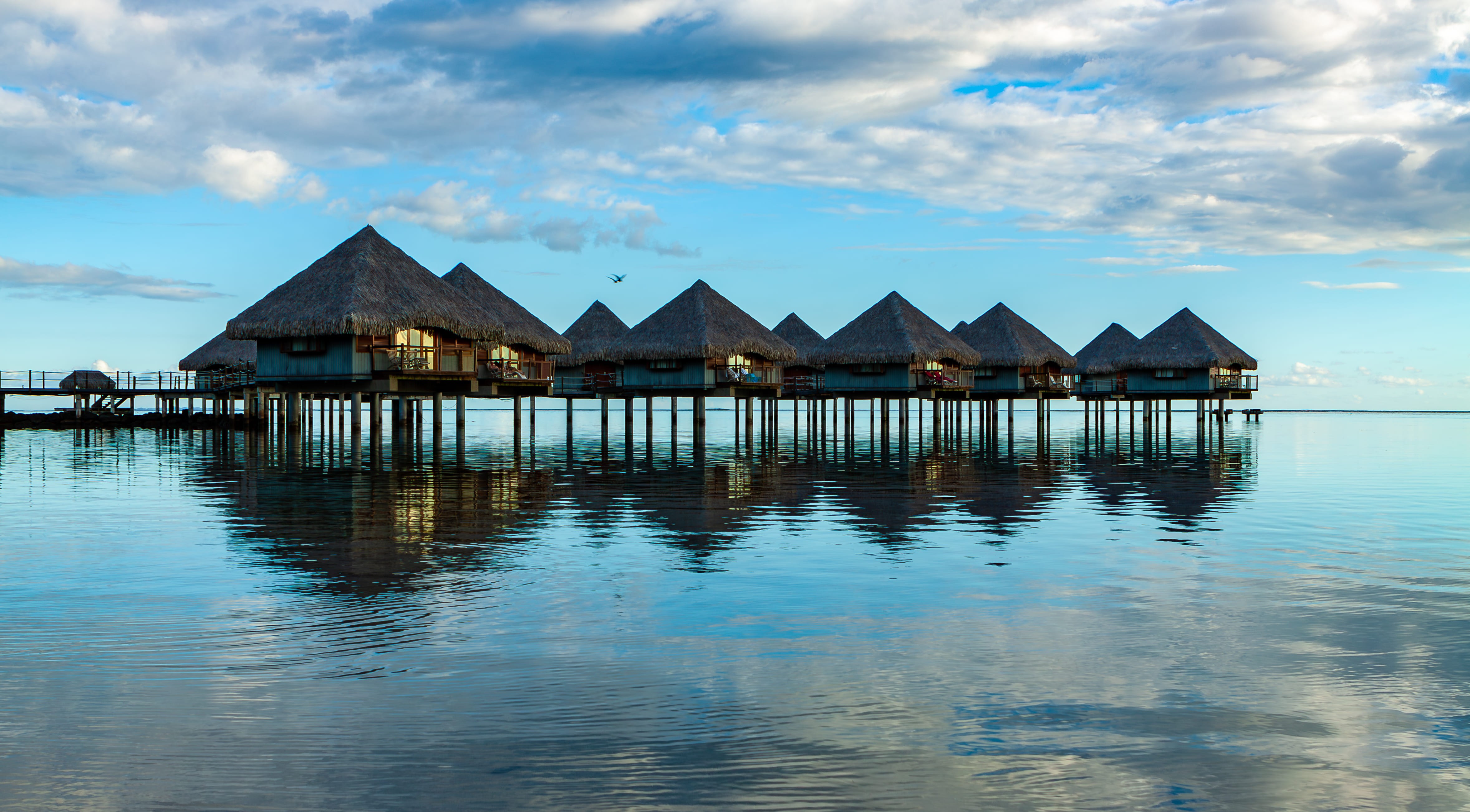 A serene lagoon with overwater bungalows reflecting in calm water under a blue sky with scattered clouds.