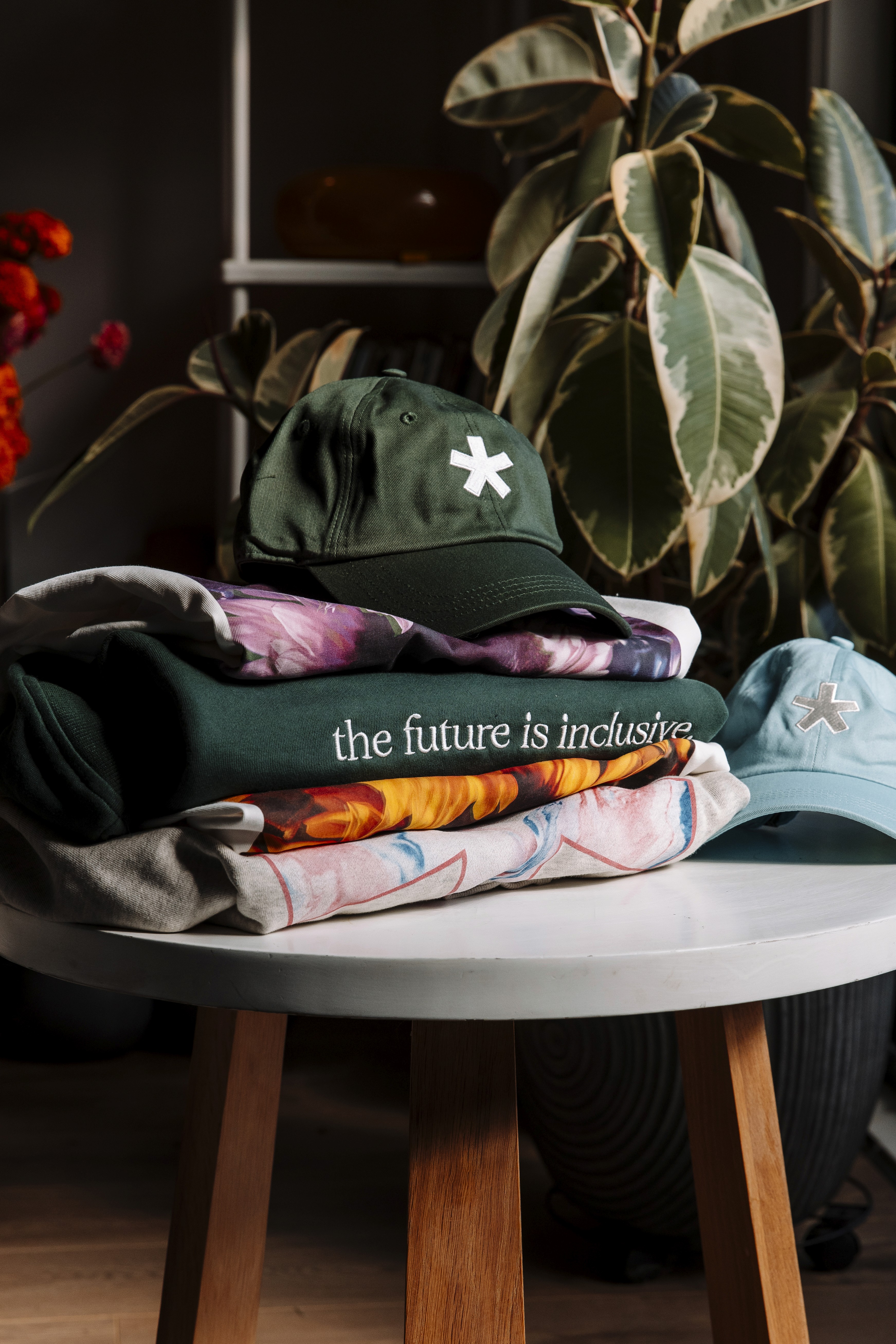 The TDS hunter green dad cap sits on top of a stack of folded clothing on a table, with plants in the background