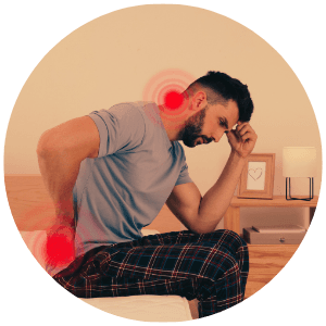 A middle aged man with dark hair and short beard, sitting on the side of his bed, leaning forward, holding his lower back with inflamation marks on his low back and back of neck.