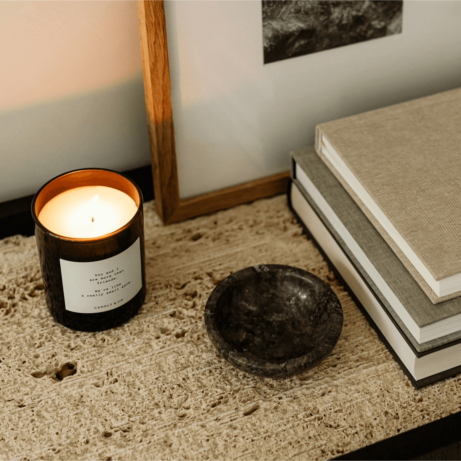 A lit candle in a dark glass holder sits on a textured stone surface next to a small black dish, with stacked books and framed monochrome art in the background, creating a warm and cozy ambiance.