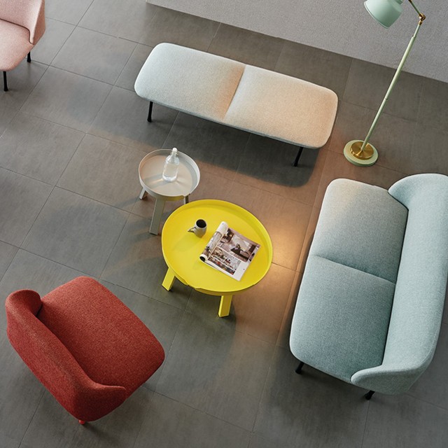 Overhead perspective of a contemporary furniture set featuring a teal fabric sofa, red lounge chair, and gray upholstered bench. Minimalist living room design with colorful nesting coffee tables and a mid-century style floor lamp on a stone tile floor.