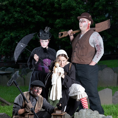 Four people in Victorian-style costumes with makeup, posing in a graveyard. One holds a shovel and another holds a doll.