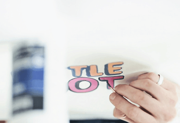 A hand holds a paintbrush, applying paint to the letters "TLE OT" on a white surface. The letters are multicolored, with "TLE" in orange, "OT" in pink, and "T" in orange. The background is blurred, with a blue and white vertical stripe visible on the left.