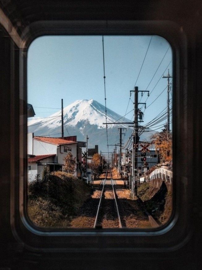 View of a railway track leading towards a snow-capped mountain, observed through a train window, with a clear blue sky.