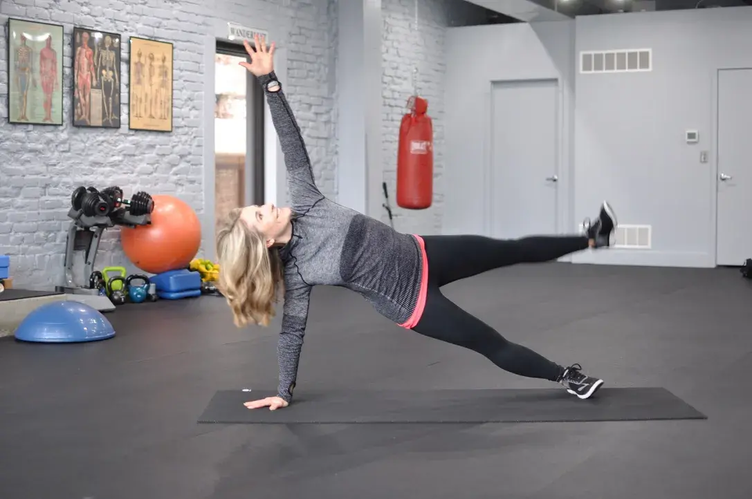 Heather Hausman exercises on a yoga mat in a gym, performing a side plank with one arm extended upwards and one leg lifted, surrounded by fitness equipment like exercise balls and weights.