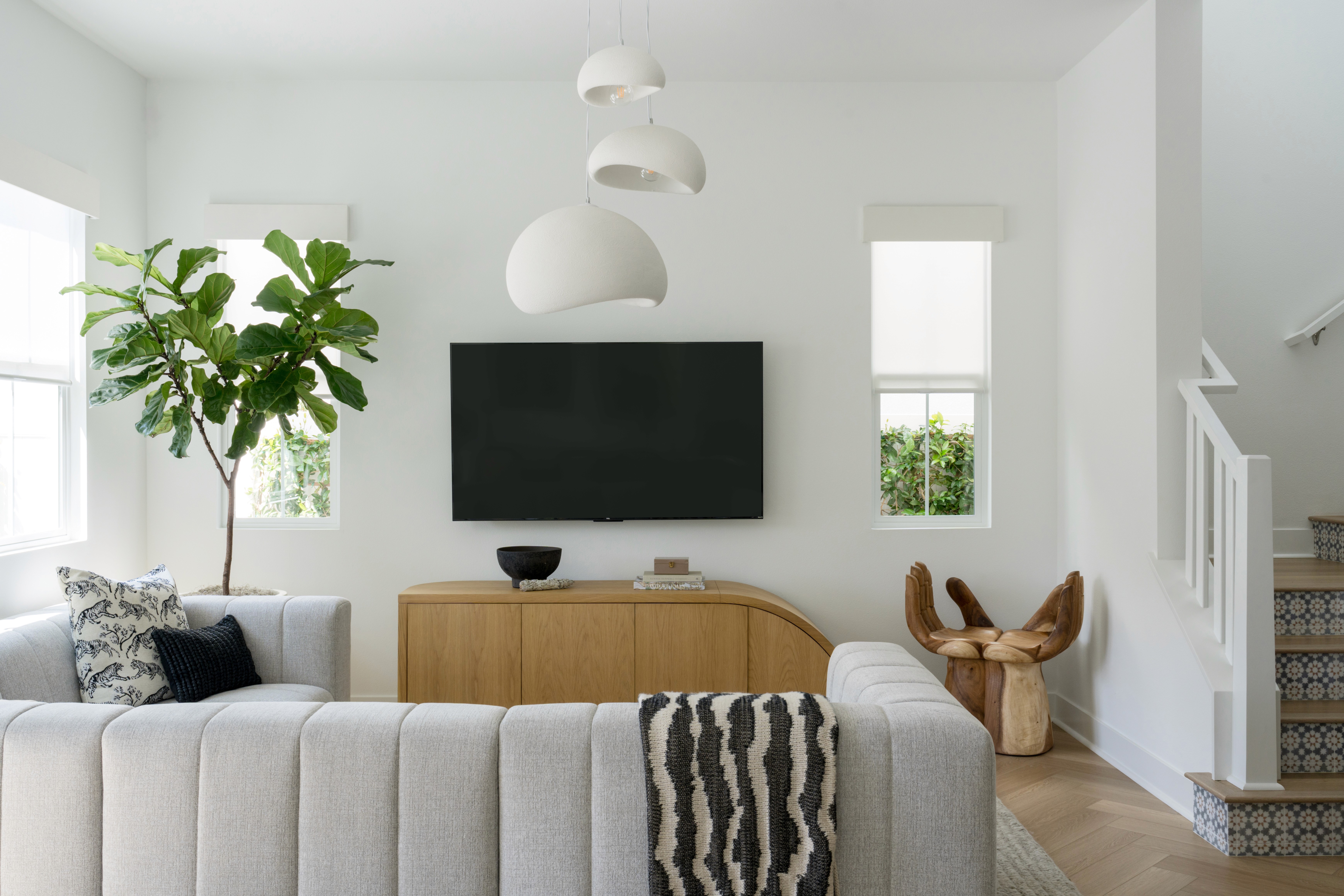 Dynamic living room with wood, curved media console, hand-shaped wood accent stool, light grey channeled sofa, and clustered white chandelier by Jubilee Interiors in Arcadia, California