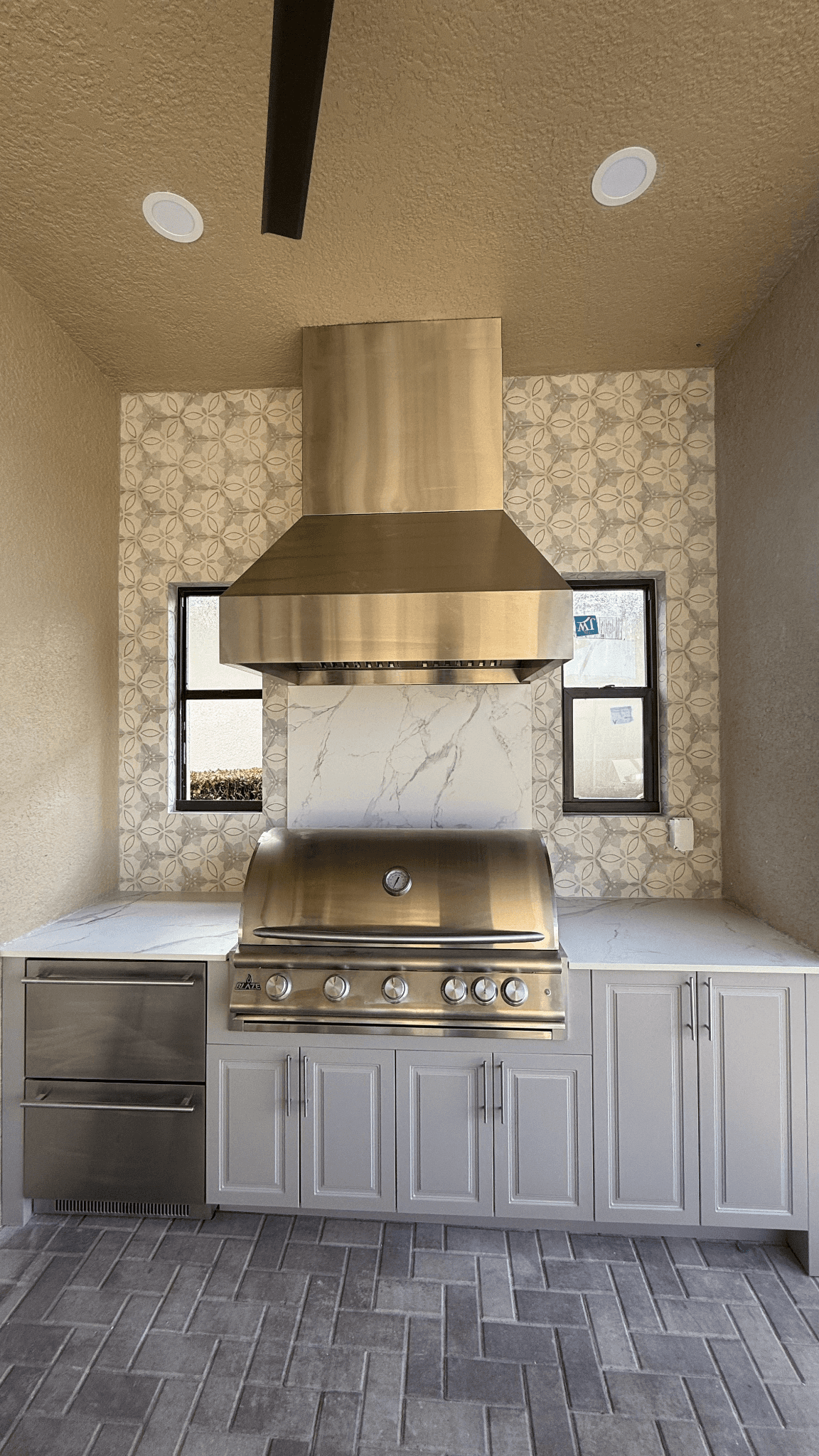 Outdoor kitchen with stainless steel grill, range hood, white cabinetry, patterned tile, and stone countertops.