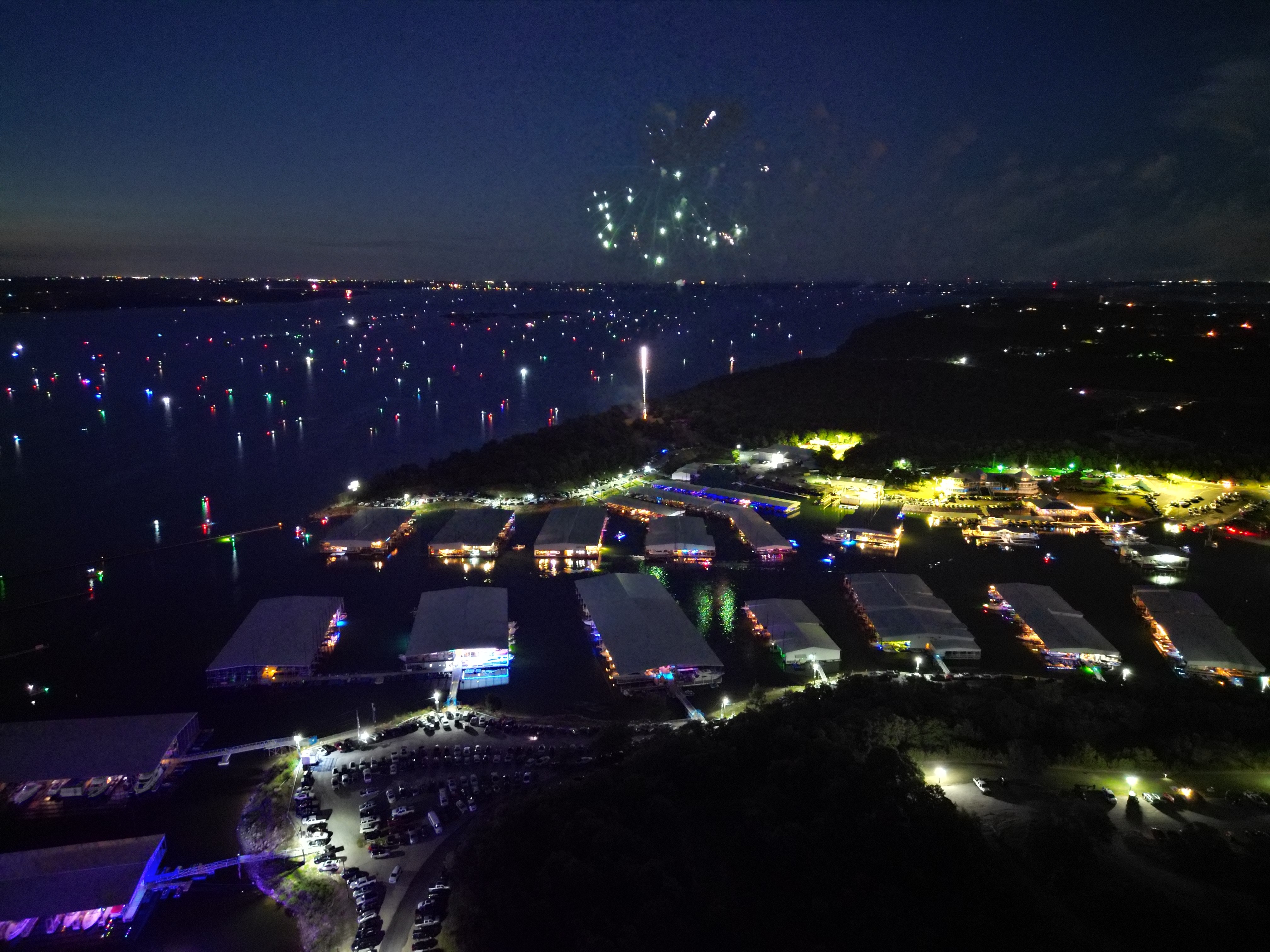 Aerial view of a vibrant marina at night, illuminated by colorful lights with numerous boats moored, and fireworks bursting in the starry sky above, creating a festive atmosphere on a calm waterbody.