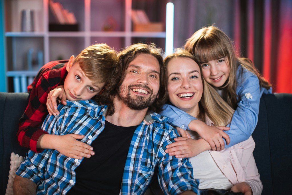 A cheerful family of four sits closely together on a couch, with the children embracing their smiling parents, showcasing their love and happiness in a warm, cozy living room setting.