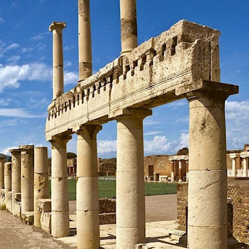 Ancient stone columns and ruins under a blue sky with scattered clouds, partial structures visible in the background.