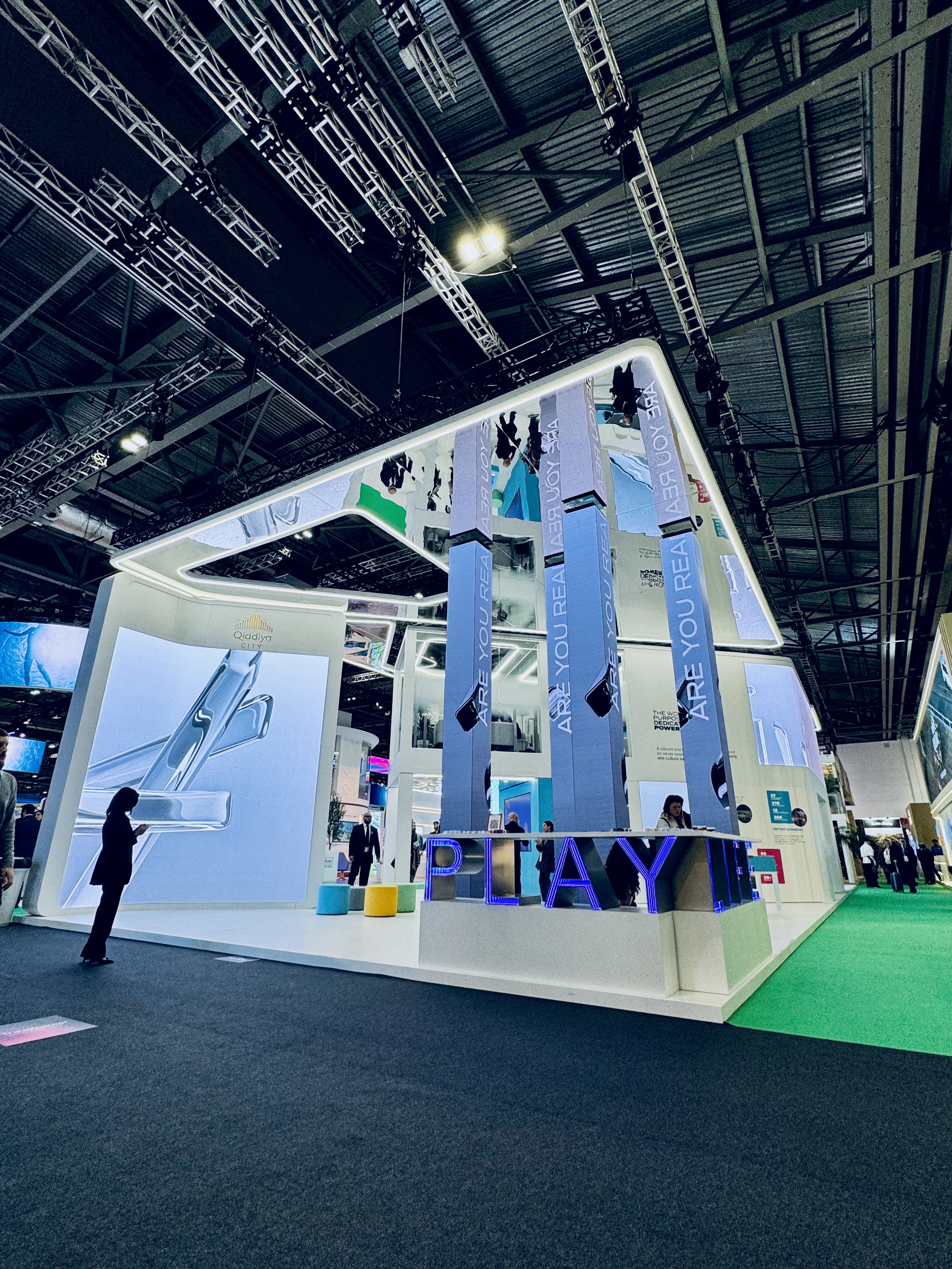 The image showcases a modern, well-lit trade show booth with tall, reflective pillars and vibrant digital displays, surrounded by attendees on a showroom floor, emphasizing innovation and design.