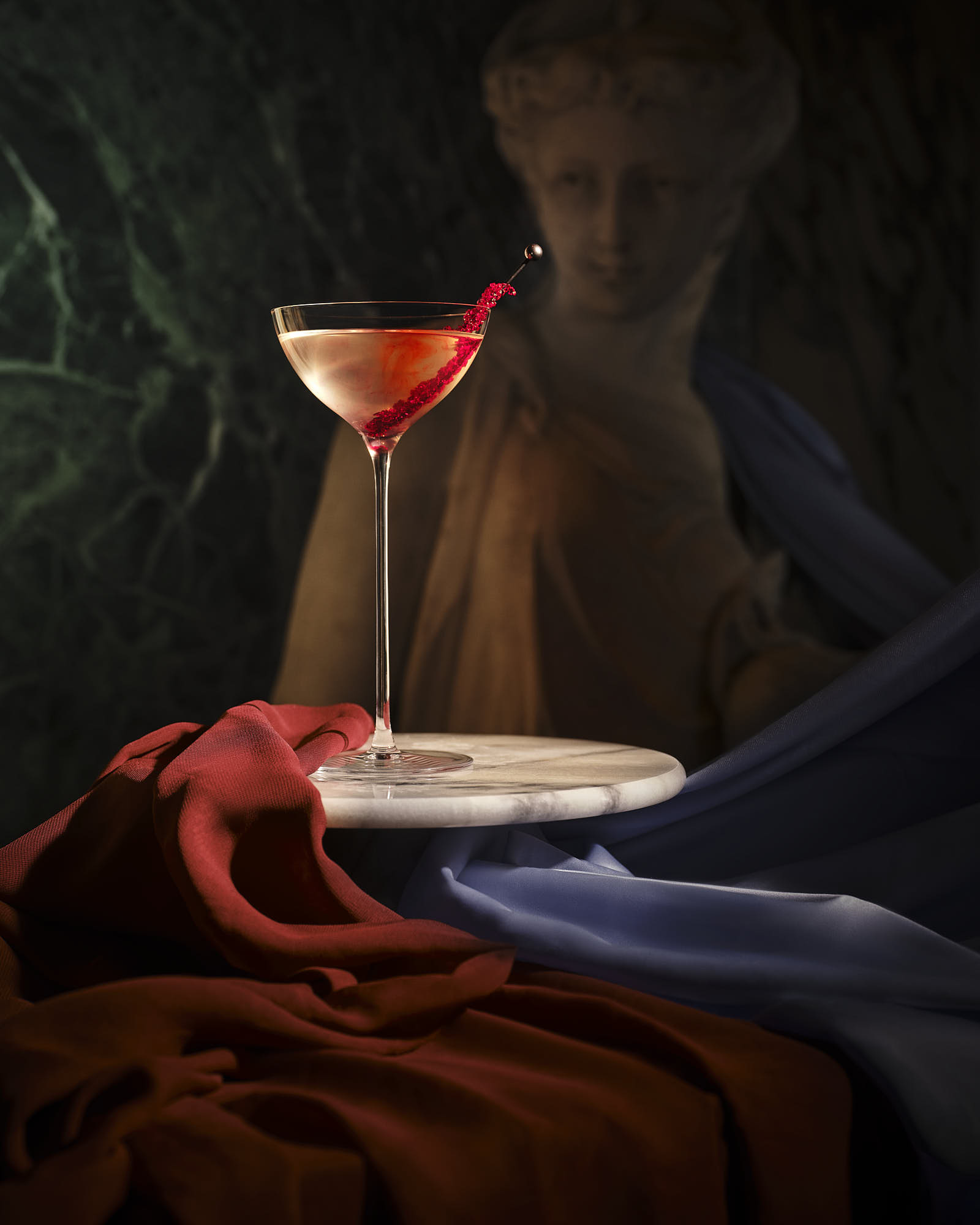 A dramatic still life featuring a cocktail glass and rich fabrics in dark surroundings.