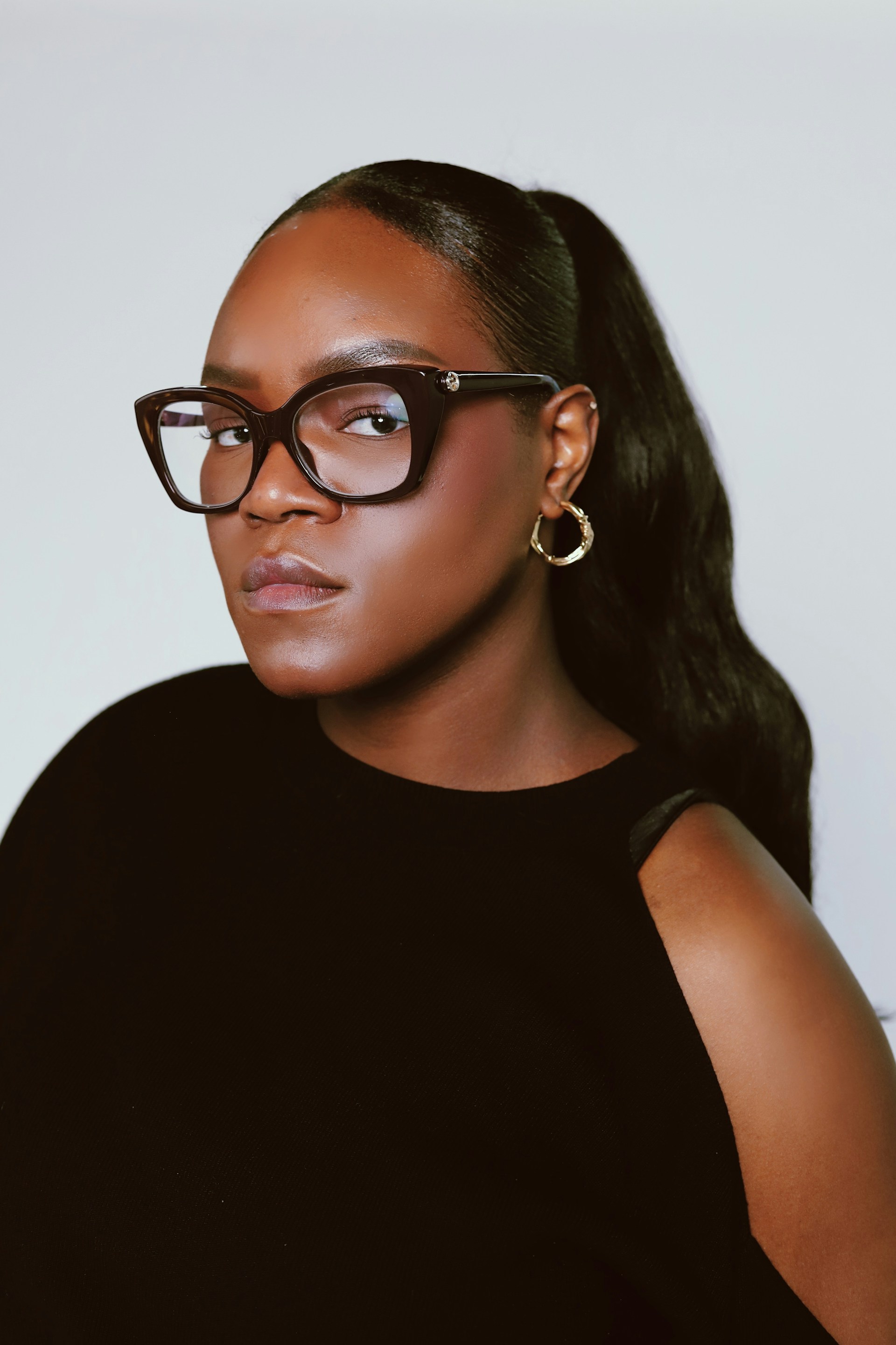 A close-up portrait of the woman wearing large, dark-framed cat-eye glasses. She has a neutral expression and is looking directly at the camera.