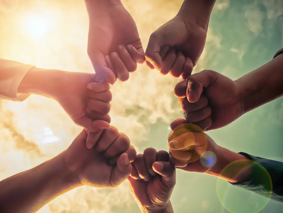 Six fists meeting in a circle, photographed from below against a bright, sun-flared sky.