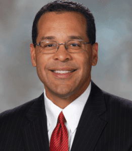 A smiling man in a suit and tie, with a light blue background.
