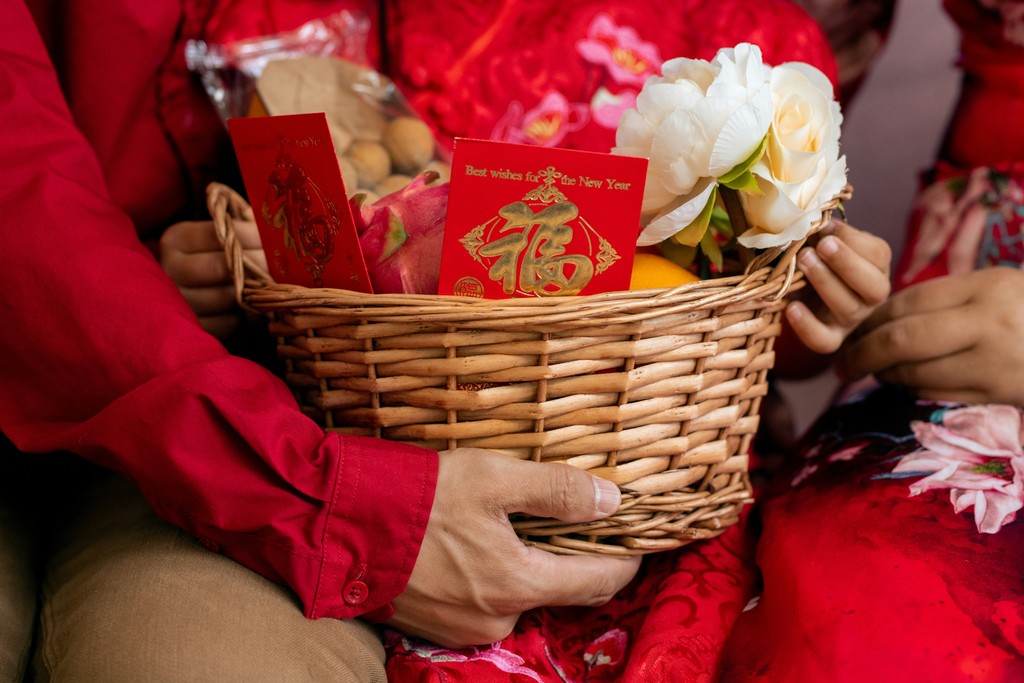 A  wicker basket filled with traditional Chinese New Year items, including red envelopes, fruits, and flowers, held by two people wearing festive red clothing, symbolizing prosperity and celebration.