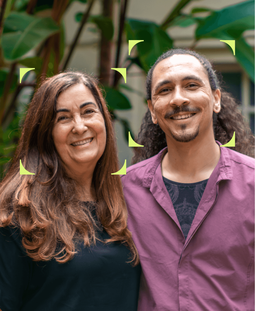 Foto de duas pessoas sorrindo lado a lado, em um ambiente externo com plantas ao fundo. À esquerda, uma mulher de cabelos longos e castanhos usa uma blusa preta; à direita, um homem de cabelos cacheados presos para trás usa uma camisa roxa. Ambos estão enquadrados por quatro marcadores gráficos em cantos arredondados, como se estivessem em destaque na imagem.