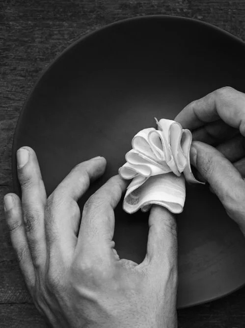 Hands carefully folding a white linen napkin into a decorative shape on a dark dinner plate, showcasing bespoke in-flight dining presentation
