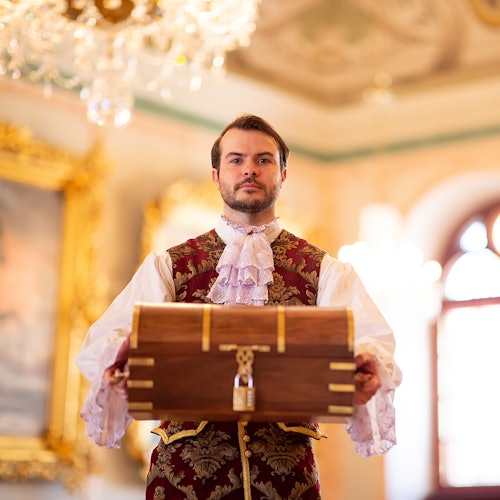 身着古装的男子在一个装饰华丽的房间里拿着一个木箱，房间内有吊灯和装裱精美的艺术品。