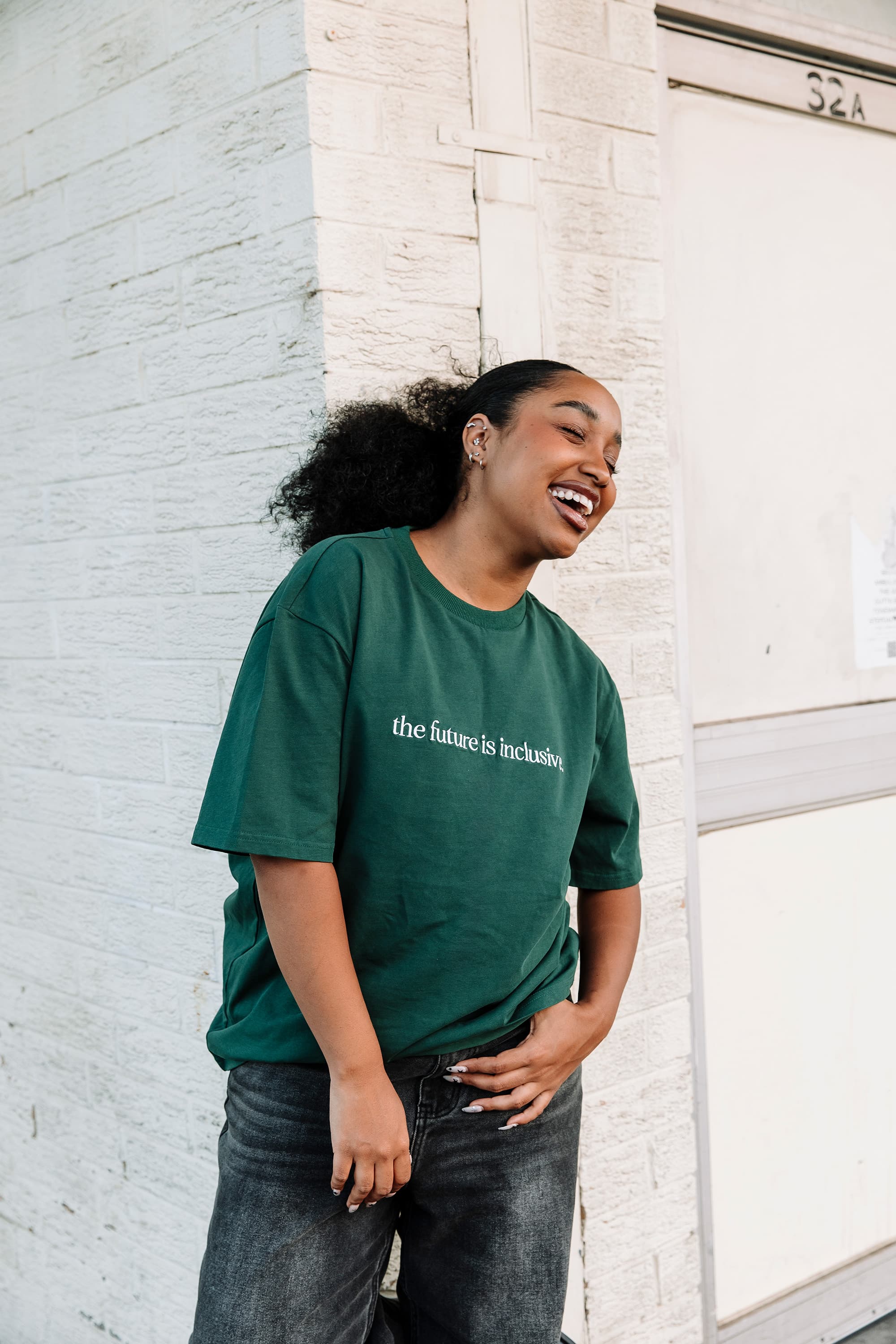 A young woman wearing a hunter green t-shirt that says 'the future is inclusive', standing in front of a white brick wall