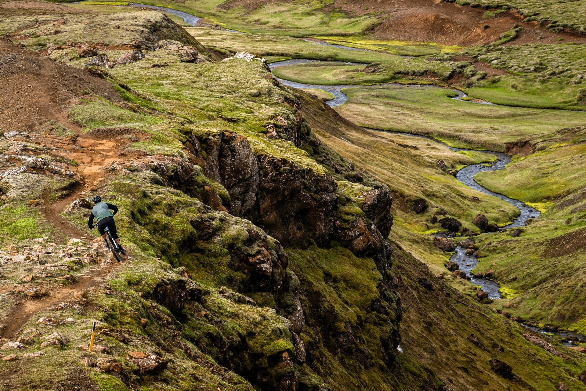 Mountain biker riding a trail above a winding river through mossy hills.