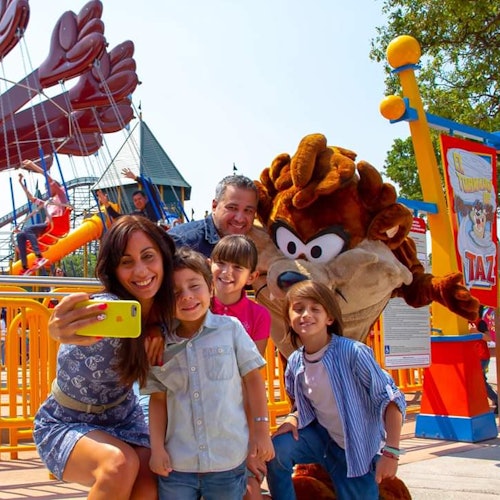 一家人正在遊樂園與一個穿著角色服裝的角色自拍，背景中可見色彩繽紛的遊樂設施和景點。