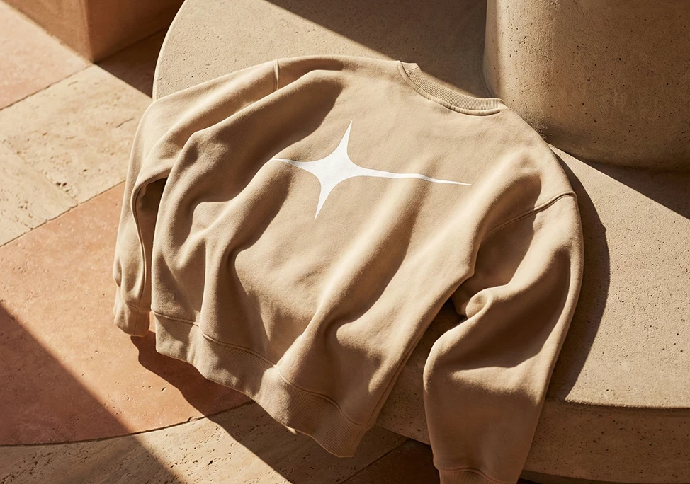 A sand-coloured sweatshirt draped over a stone ledge in warm sunlight, printed with the white Action Replay star.