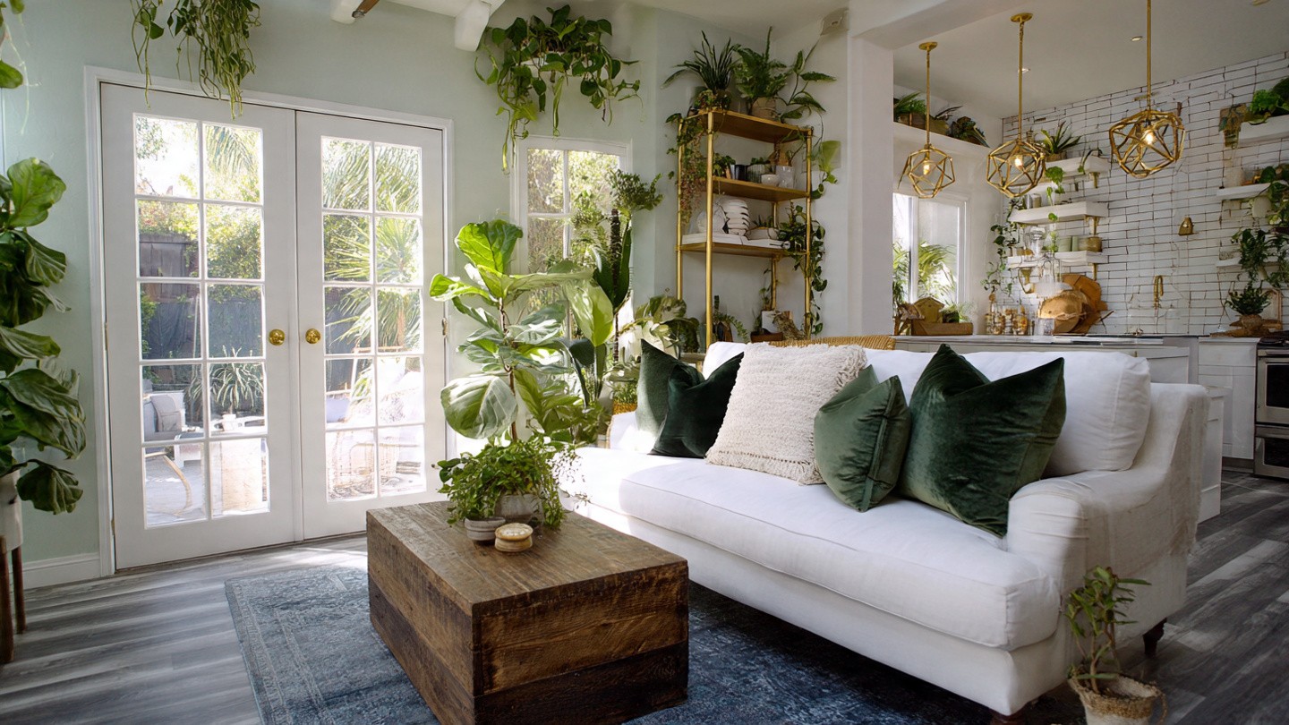 Bright living room with white sofa adorned by green cushions, surrounded by lush plants. Sunlight streams through French doors, illuminating the cozy, modern space.