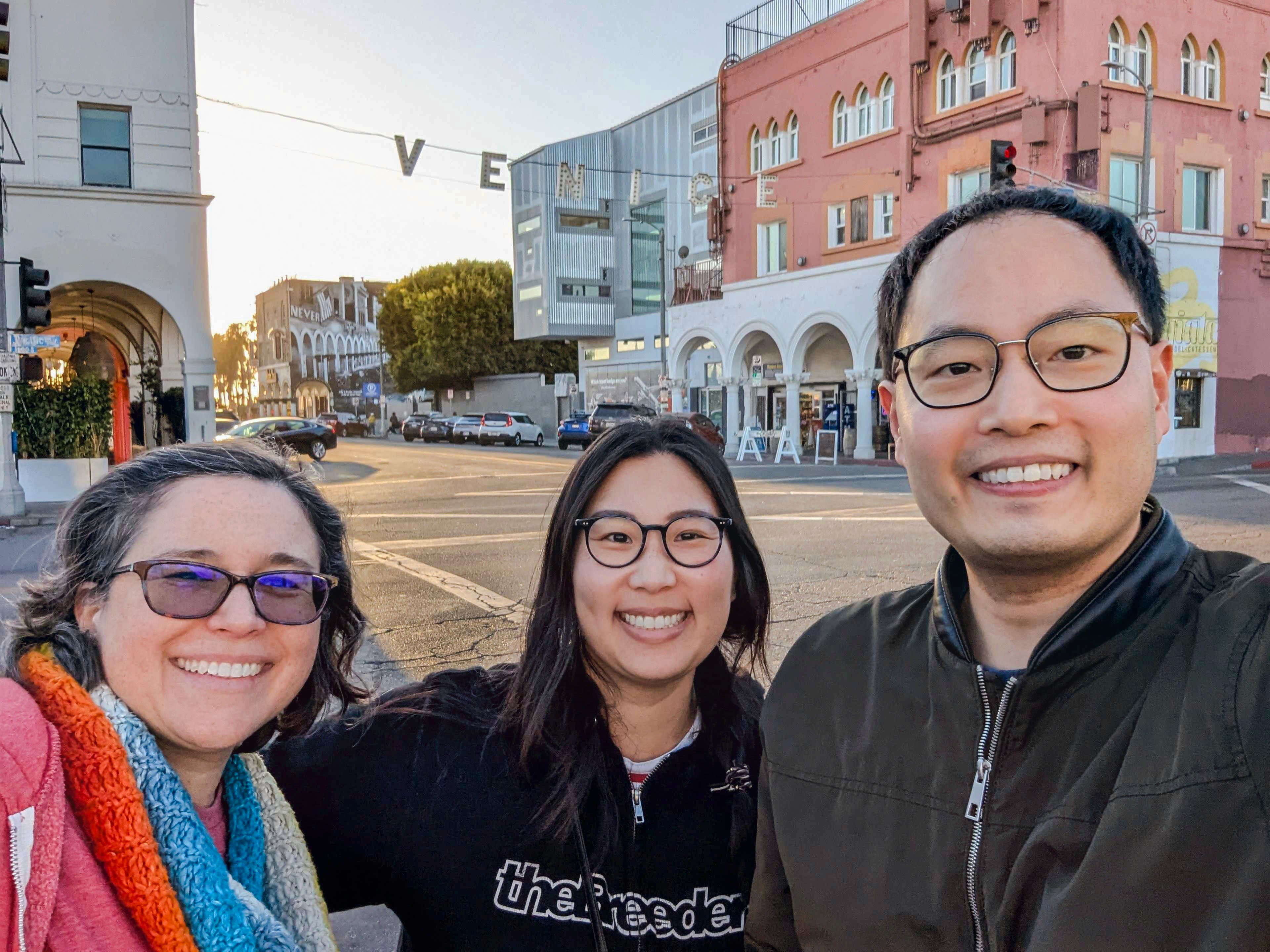 Three people smiling in front of the Venice sign in Venice Beach, California, with colorful buildings and sunset light in the background.