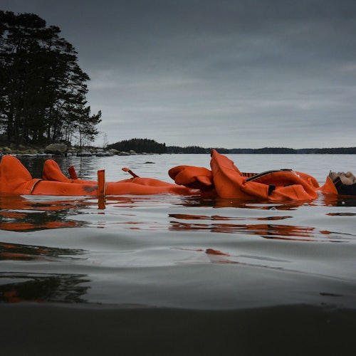 Two people in orange life suits float on a calm body of water, with a cloudy sky and tree-lined shore in the background.