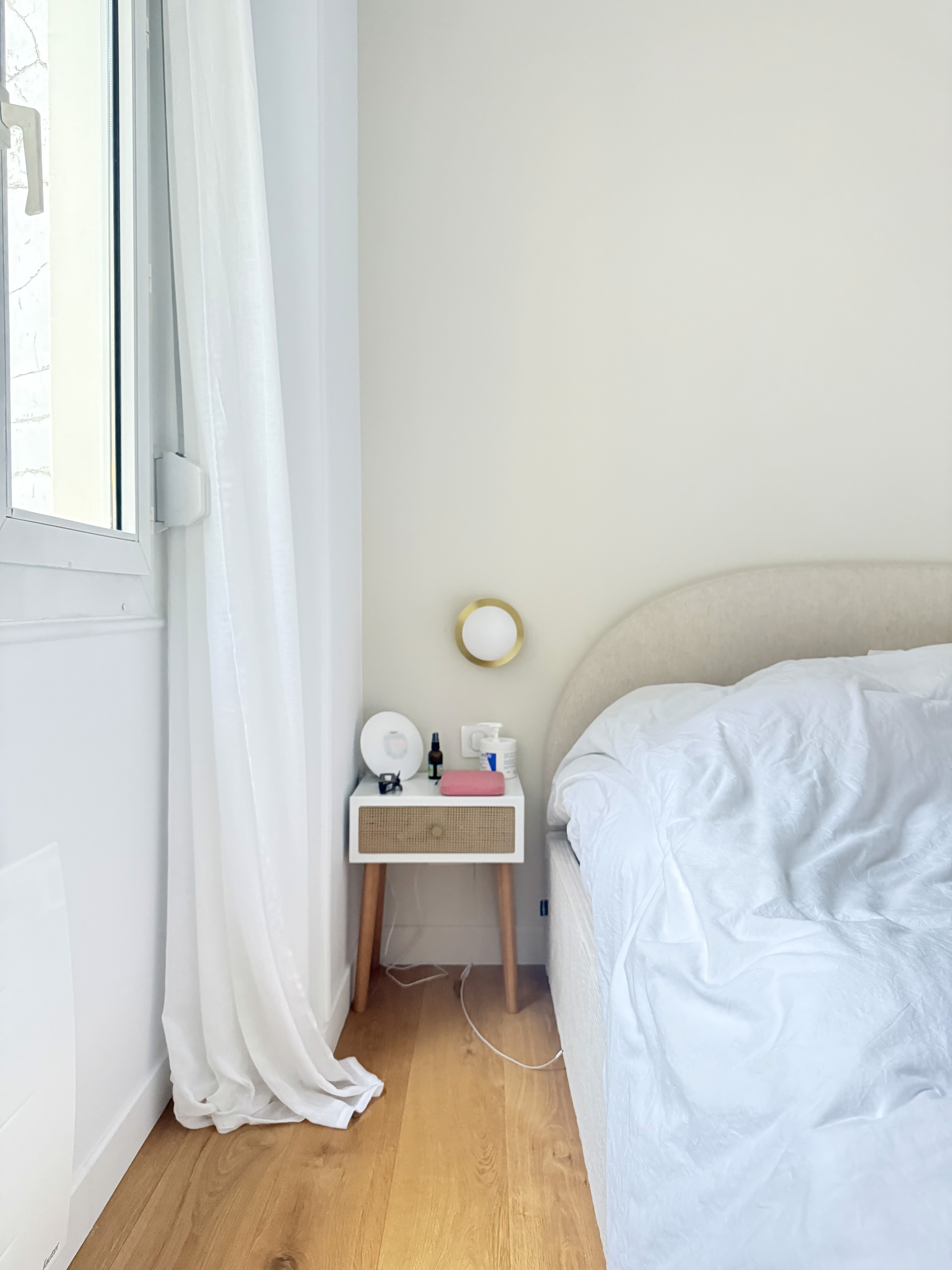 Détail de la chambre principale avec table de chevet design, linge de lit blanc et petite applique murale dorée.