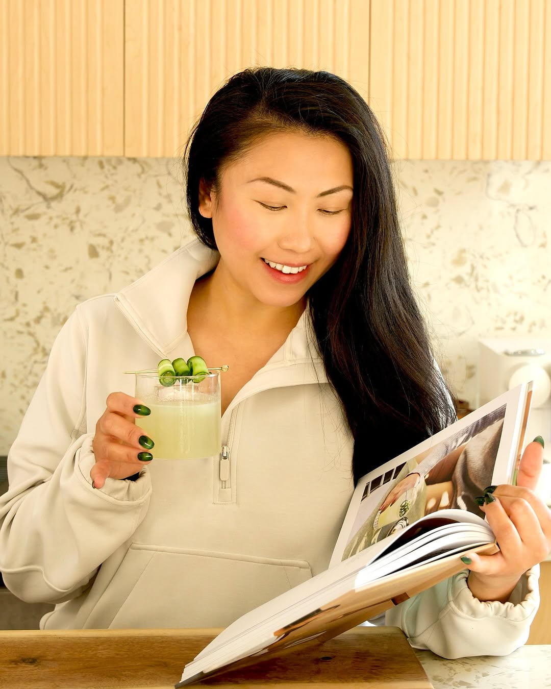 A relaxed moment at Node Studio, featuring a woman enjoying a fresh cucumber cocktail while browsing a book, with the warm tones of the studio’s semi-equipped kitchen in the background