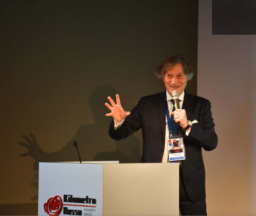 Relatore in abito scuro durante una conferenza al Kilometro Rosso, mentre parla al microfono e gesticola dal podio.