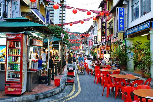 Busy street in Asia reminiscent of Japan, featuring a vibrant market scene with red tables lining the street where people leisurely enjoy their meals.