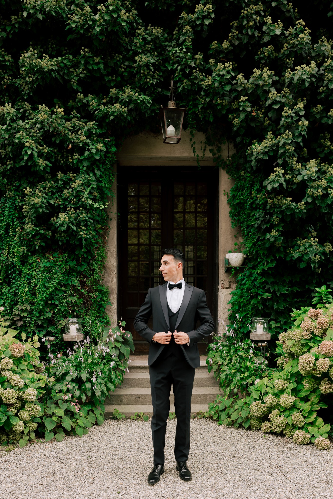 Sposo in smoking davanti all'ingresso di Villa Ferrari circondato da edera e fiori bianchi, reportage matrimoniale