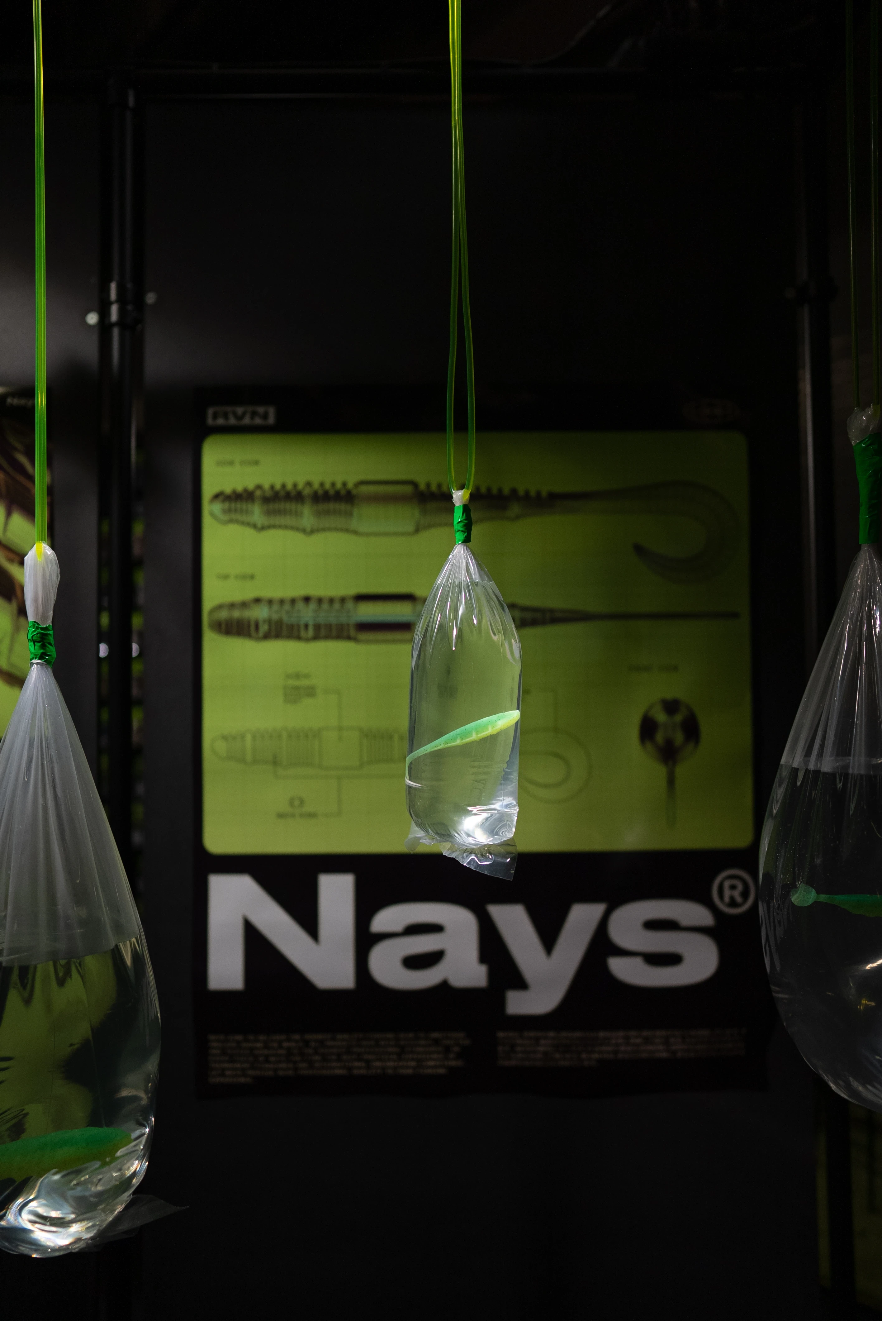 Hanging water bag with green soft-bait lure in front of a technical Nays product poster in acid green.