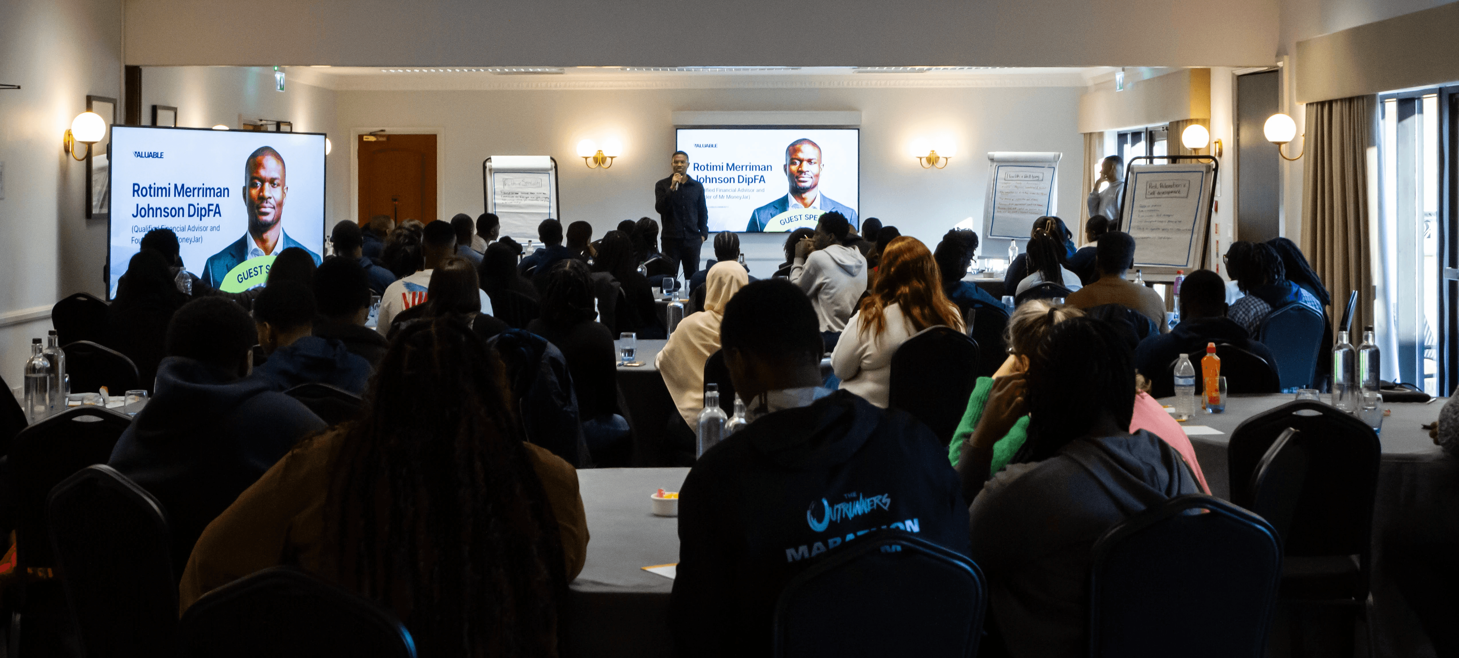 A group of people seated in a conference room, facing a speaker at the front. Natural light from windows.