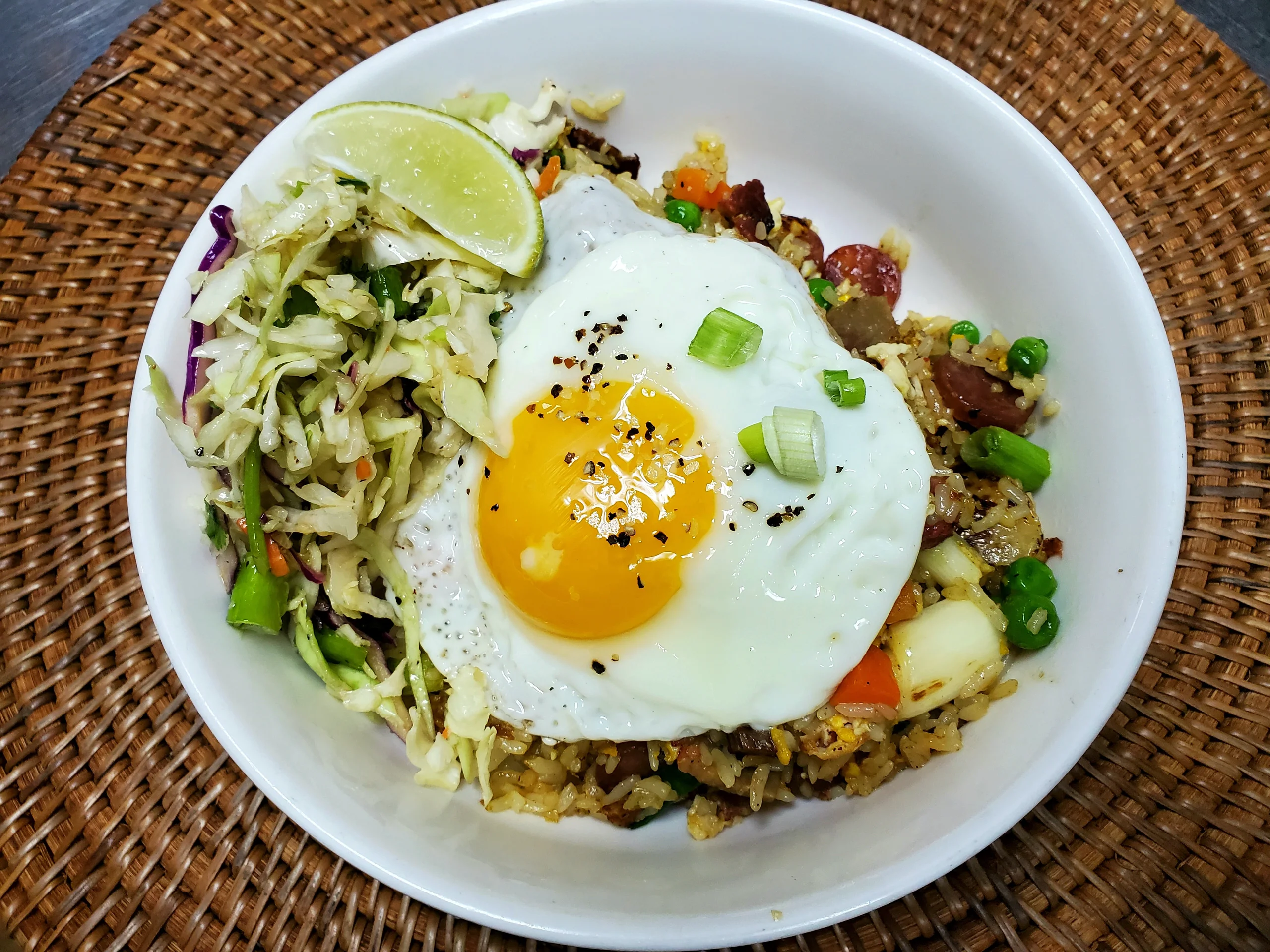 A white bowl featuring a mound of fried rice with peas, carrots, and pineapple, topped with a large sunny-side-up fried egg and served with a side of cabbage slaw and a lime wedge.