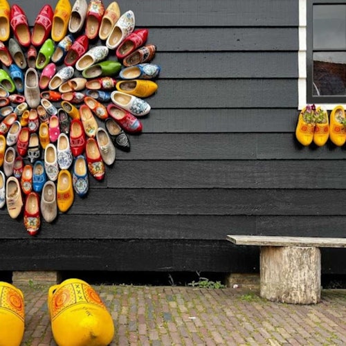 Wooden clogs arranged in a heart pattern on a black wooden wall, with a window and bench below. Large yellow clogs are on the ground.