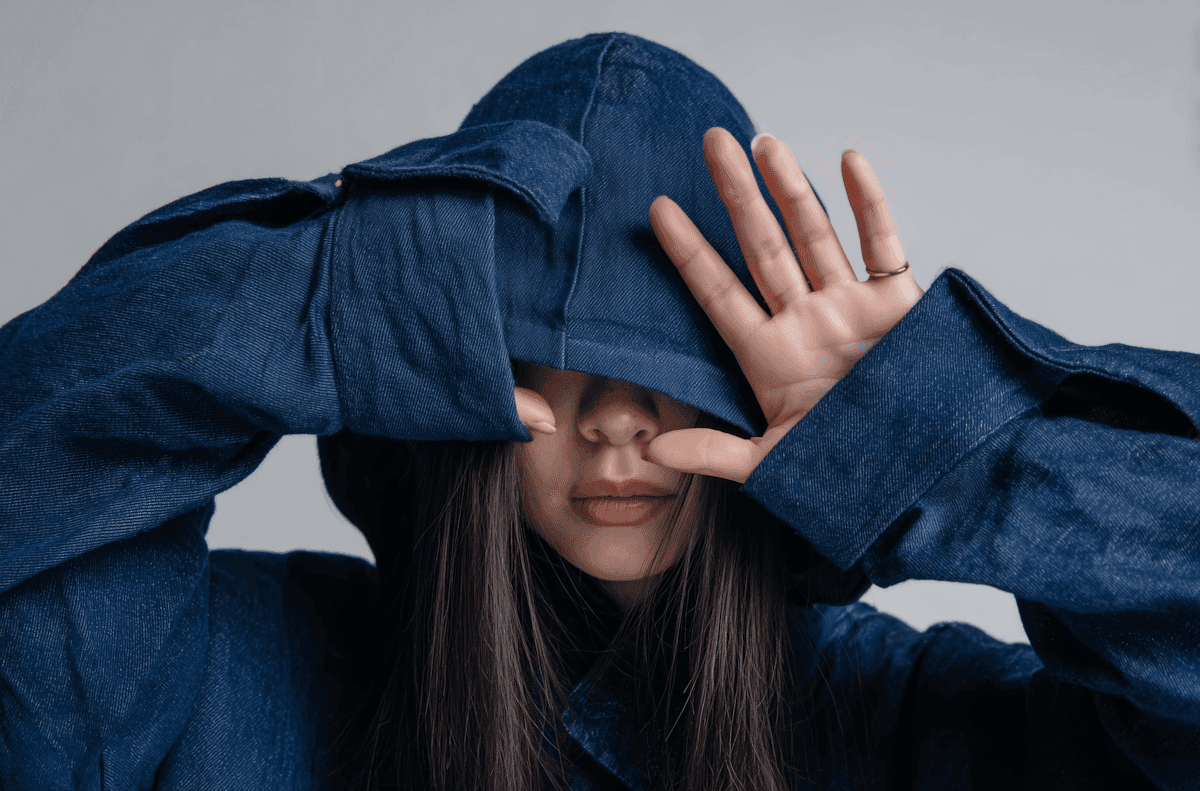 A person wearing a dark blue denim jacket covers their face with the hood and sleeves, against a simple, light gray background.