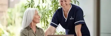 A smiling Florence nurse in navy uniform standing beside an older woman seated at a desk with a laptop, both engaged in a warm interaction