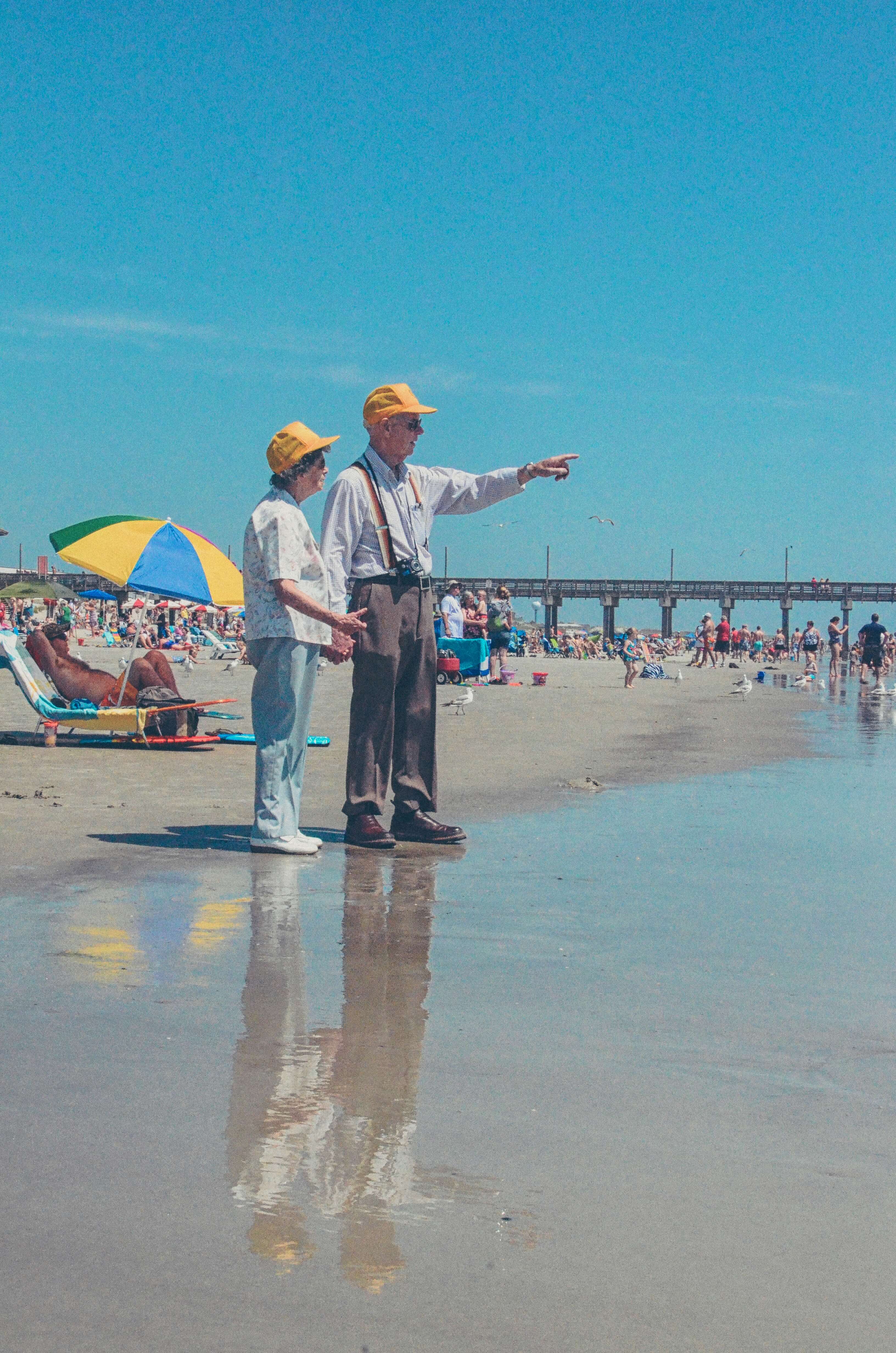 an elderly couple in yellow trucker hats on beach pointing out to the water