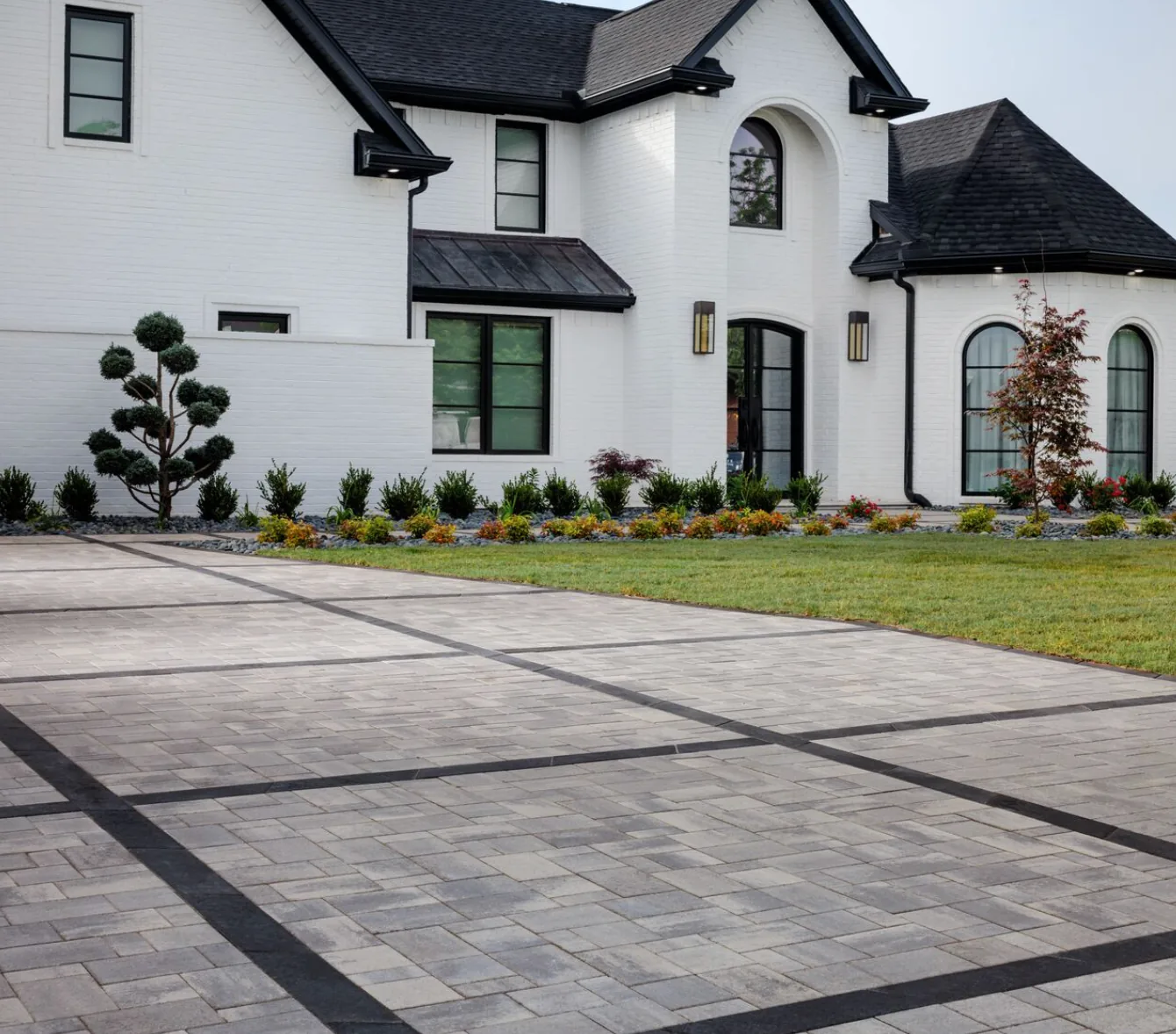 Gray paver driveway divided by dark bands in front of a white two-story house.