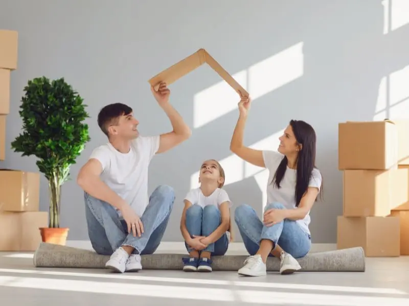 A family of three sits on the floor of a room filled with moving boxes, symbolizing a new beginning in a new home. The father and mother, both in casual white shirts and jeans, are playfully holding a picture frame above their heads to form a makeshift house, with their child sitting between them looking up, smiling at the imaginary roof. A potted plant adds a touch of greenery to the scene, suggesting a warm and loving household environment.