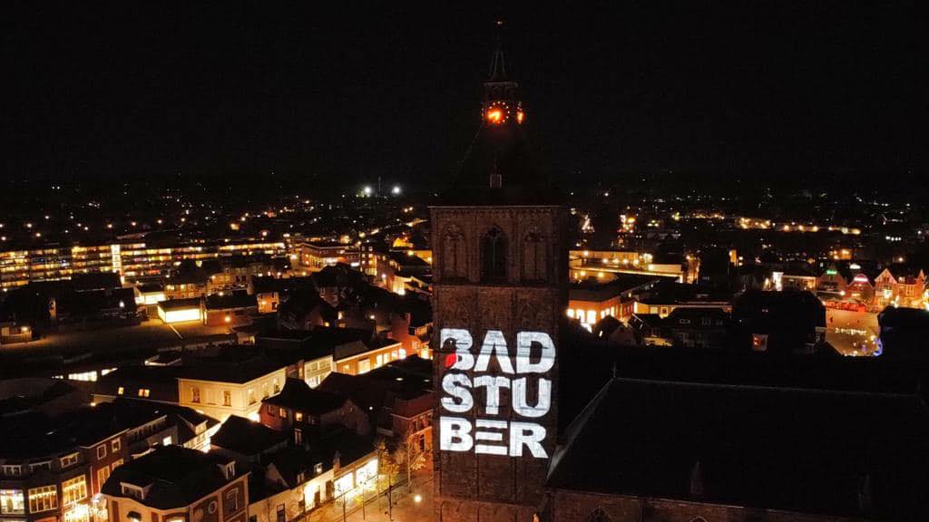 Het Badstuber-logo geprojecteerd op de kerktoren van Oldenzaal, 's avonds van bovenaf gezien