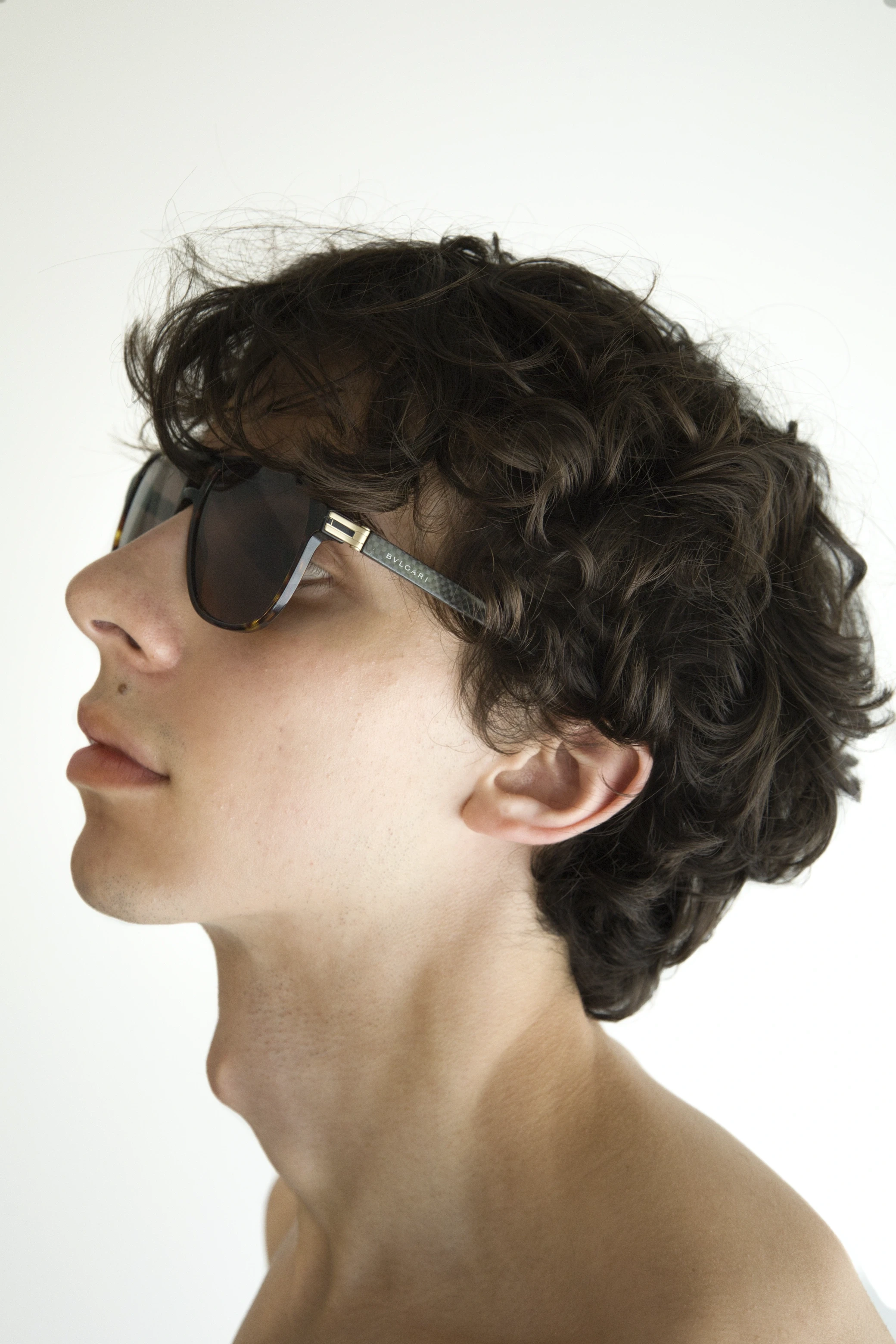 A person with curly hair wearing sunglasses, gazing sideways against a plain white background.
