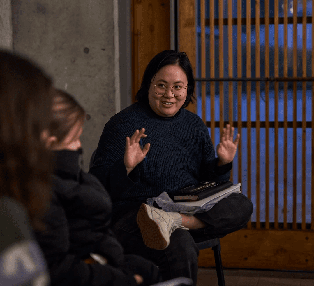 A person sits cross-legged on a chair in a small group discussion, speaking with their hands raised mid-gesture. They wear glasses and a dark jumper, with notebooks on their lap. Several people sit around them listening. The setting appears informal, with concrete walls and wooden slats behind them, suggesting a workshop or creative discussion space in the evening.